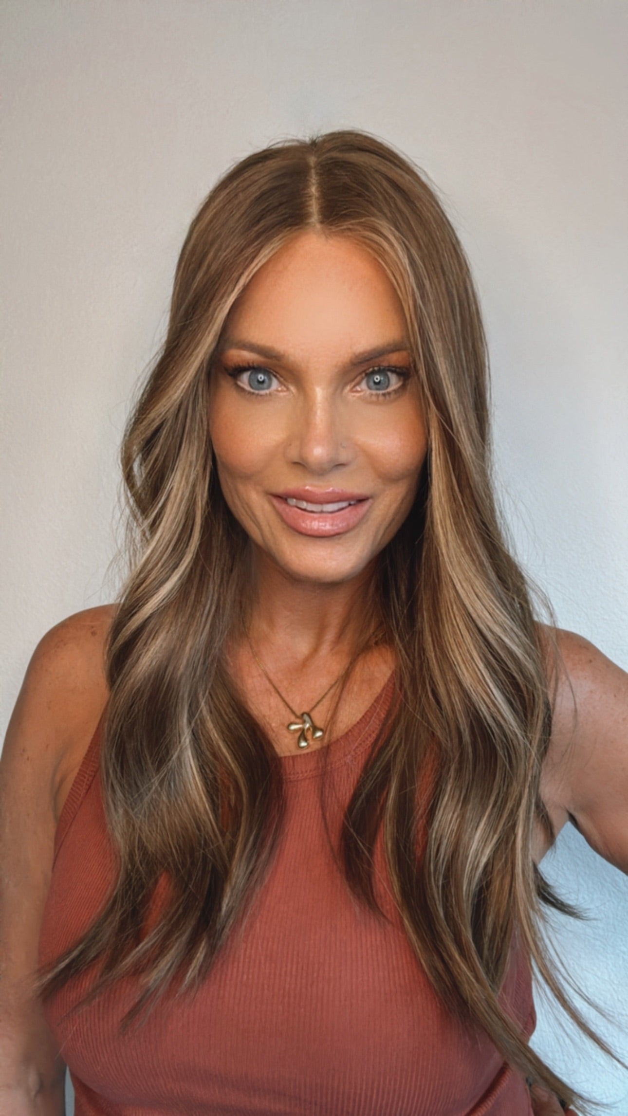Woman with long, wavy human hair wig wearing a rust-colored top against a plain background